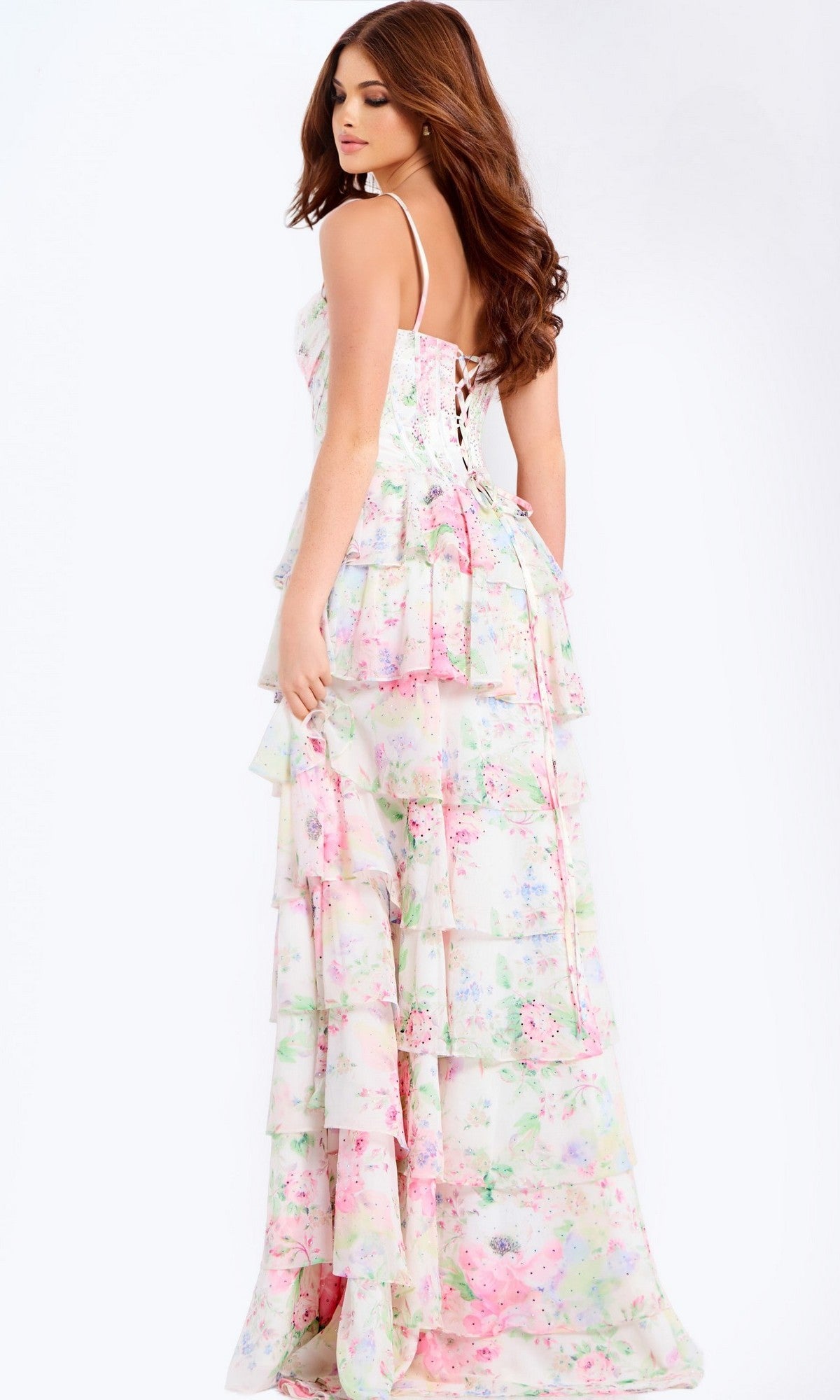 Woman models the Jovani 47282 long prom dress—a tiered, floral pastel maxi with thin straps, corset bodice, and lace-up back—while standing against a plain background.